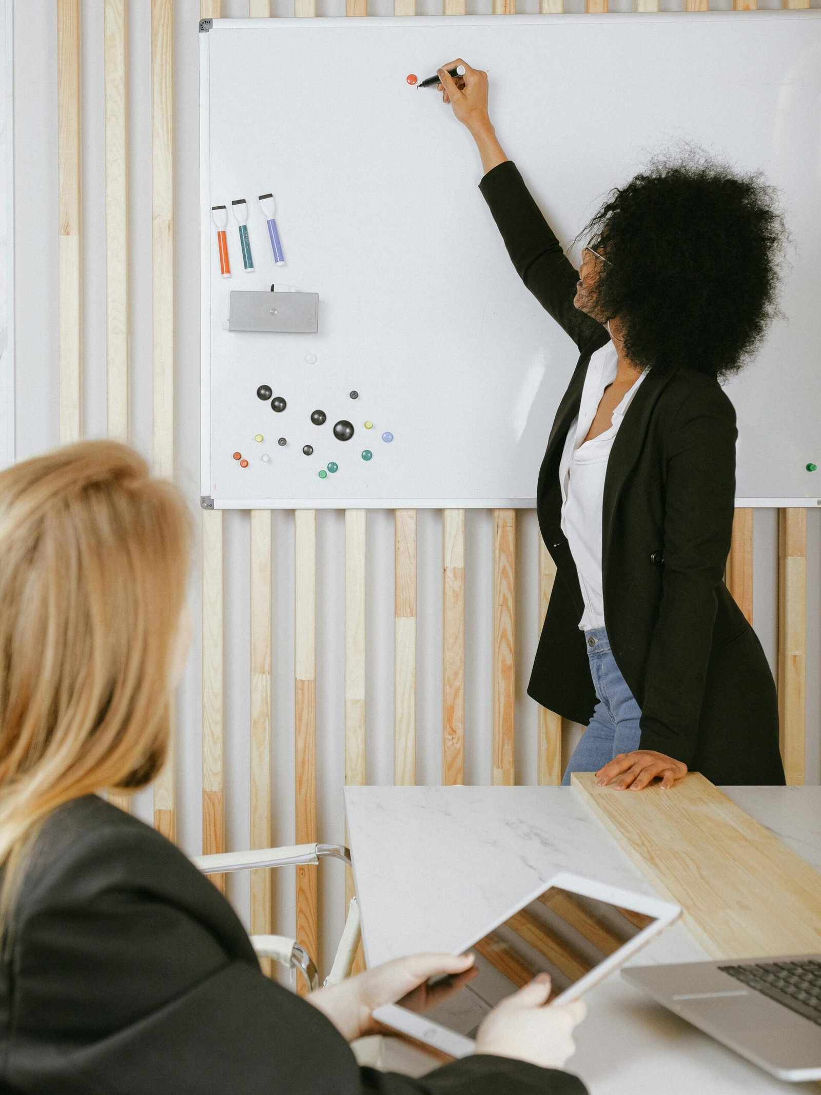 Two women engaged in a business presentation with a whiteboard in a modern office setting.